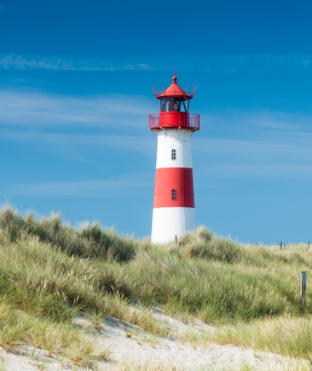 Ein rot-weiß gestreifter Leuchtturm mit roter Laterne steht hinter einer mit Dünengras bewachsenen Sanddüne Ein rot-weiß gestreifter Leuchtturm mit roter Laterne steht hinter einer mit Dünengras bewachsenen Sanddüne