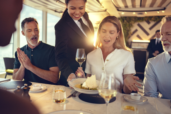 Eine Kellnerin serviert Gästen in einem eleganten Schiffsrestaurant ein Gericht, während die Gruppe lächelnd am Tisch sitzt.